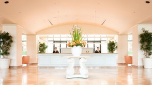 Un área de recepción con un mostrador blanco, dos recepcionistas y un jarrón grande de flores.