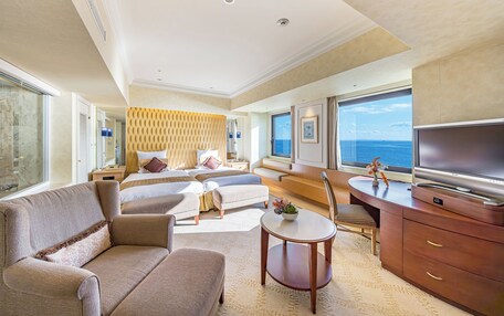 A spacious hotel room with a large bed, a sofa, a desk with a TV, and a view of the sea.