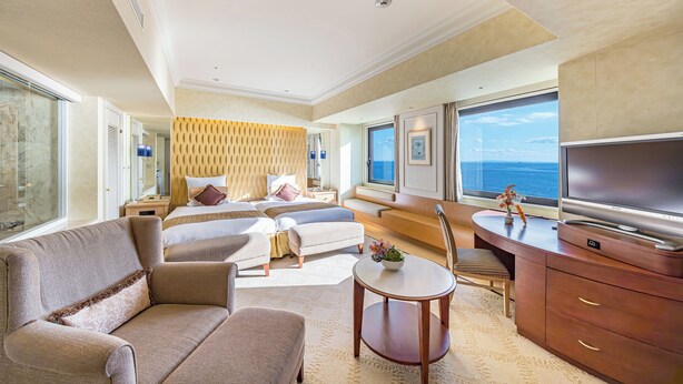 Amplia habitación de hotel con una cama grande, un sofá, un escritorio con televisor y vistas al mar.