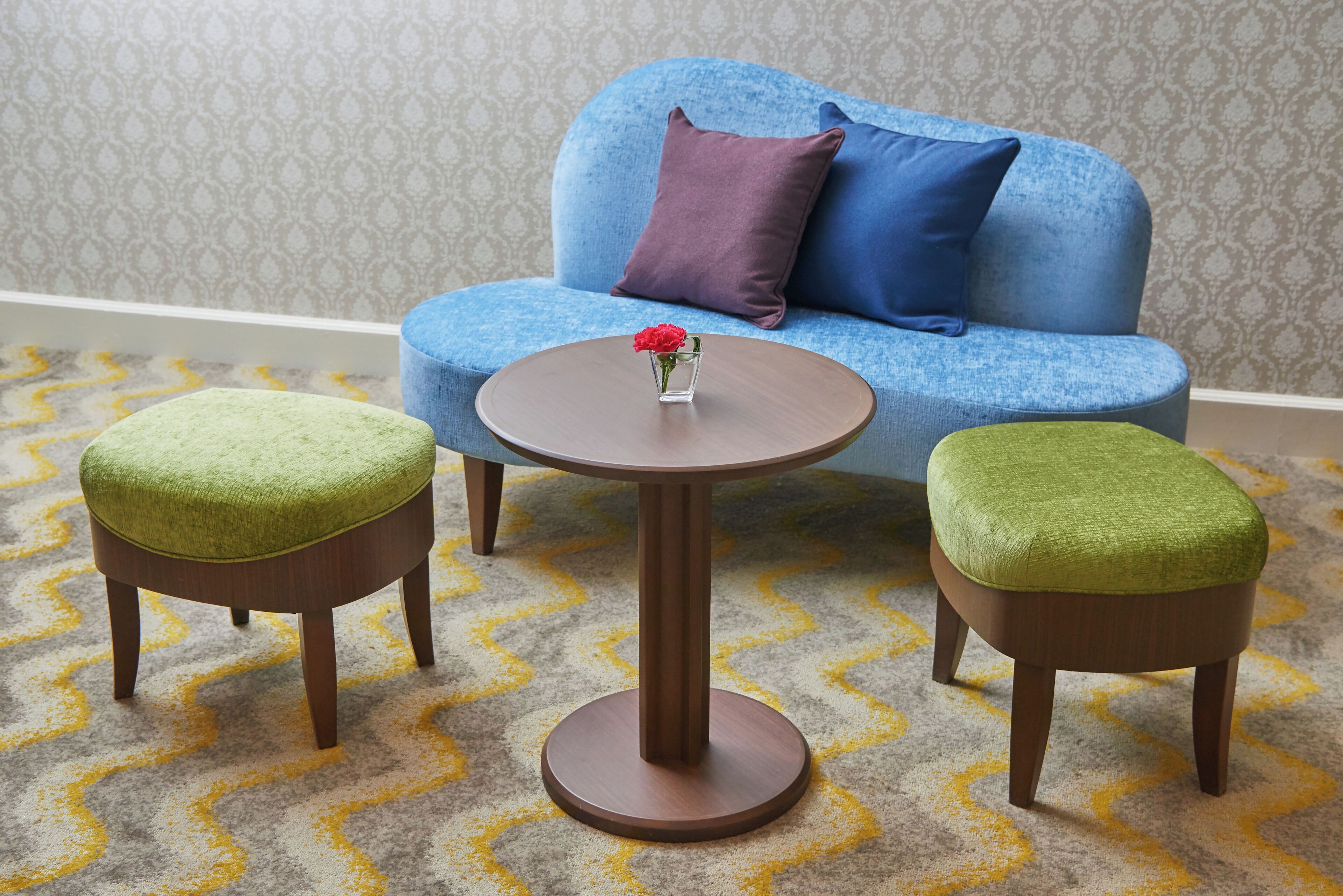 A room with a blue sofa, two green ottomans, and a round wooden table with a vase of flowers.