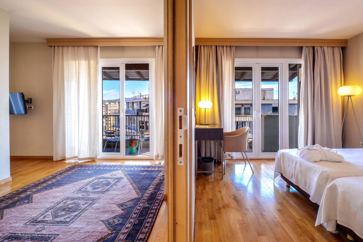 A hotel room with a large sliding glass door to a balcony, a bed, a desk with a chair, a floor lamp, and a patterned rug.