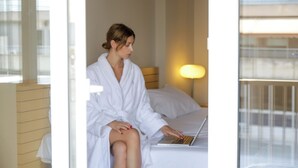 A person in a white bathrobe sitting on a bed with a laptop, in a room with a window and a bedside lamp.