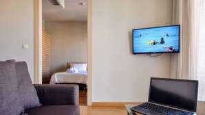 A hotel room with a sofa, a bed, a TV displaying a beach scene, and a laptop on a table.