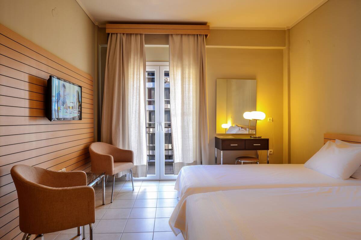 A hotel room with two beds, a desk with a lamp, a chair, a television mounted on the wall, and a view of the outside through a sliding door.