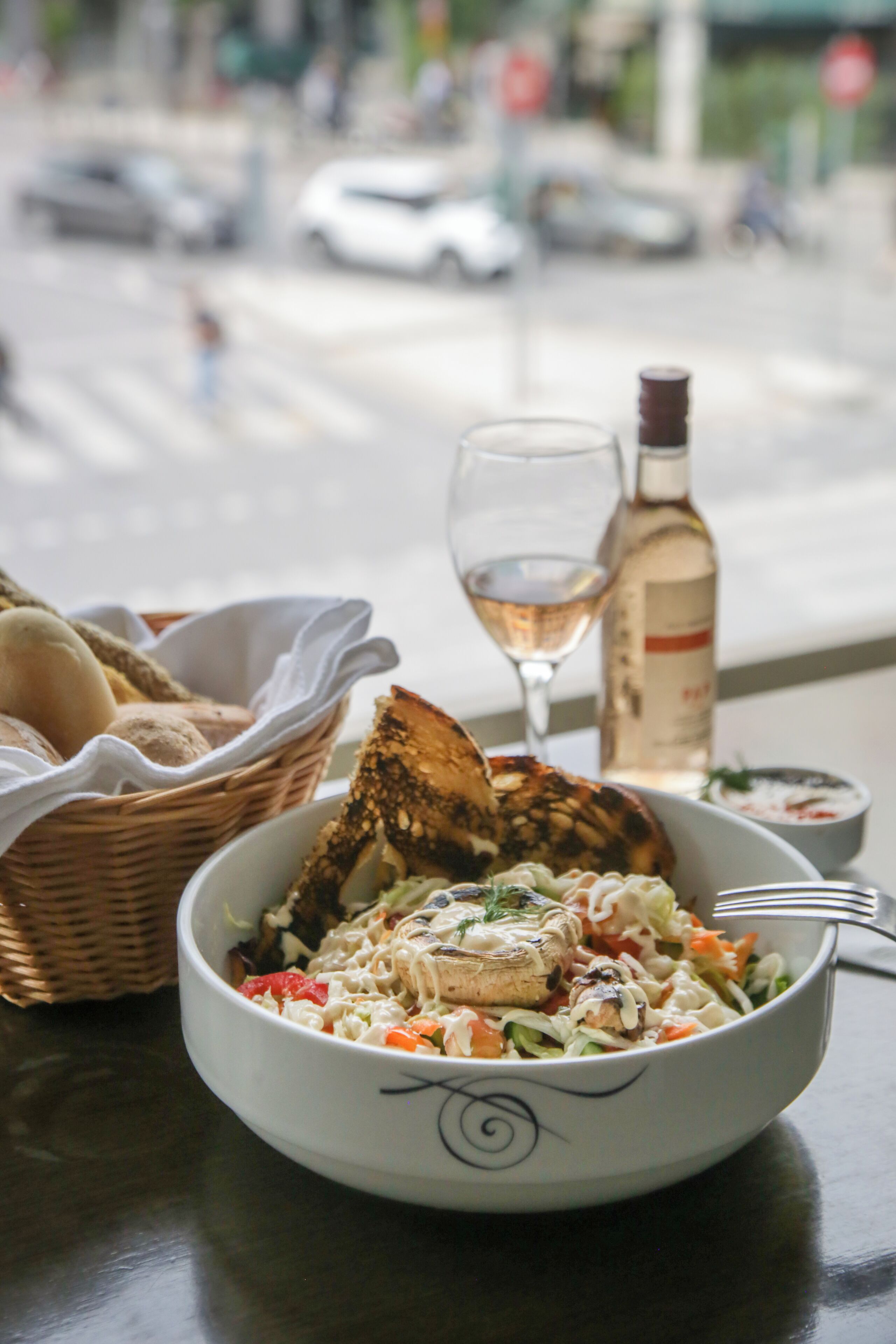 A bowl of food with bread and a bottle of wine on a table by a window.
