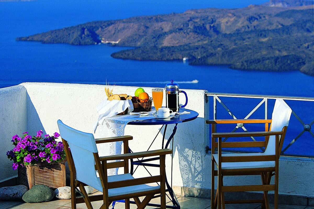 Een tafel met een koffiepot, een fruitmand en een glas sinaasappelsap op een balkon met uitzicht op een blauwe zee en een bergachtig landschap.