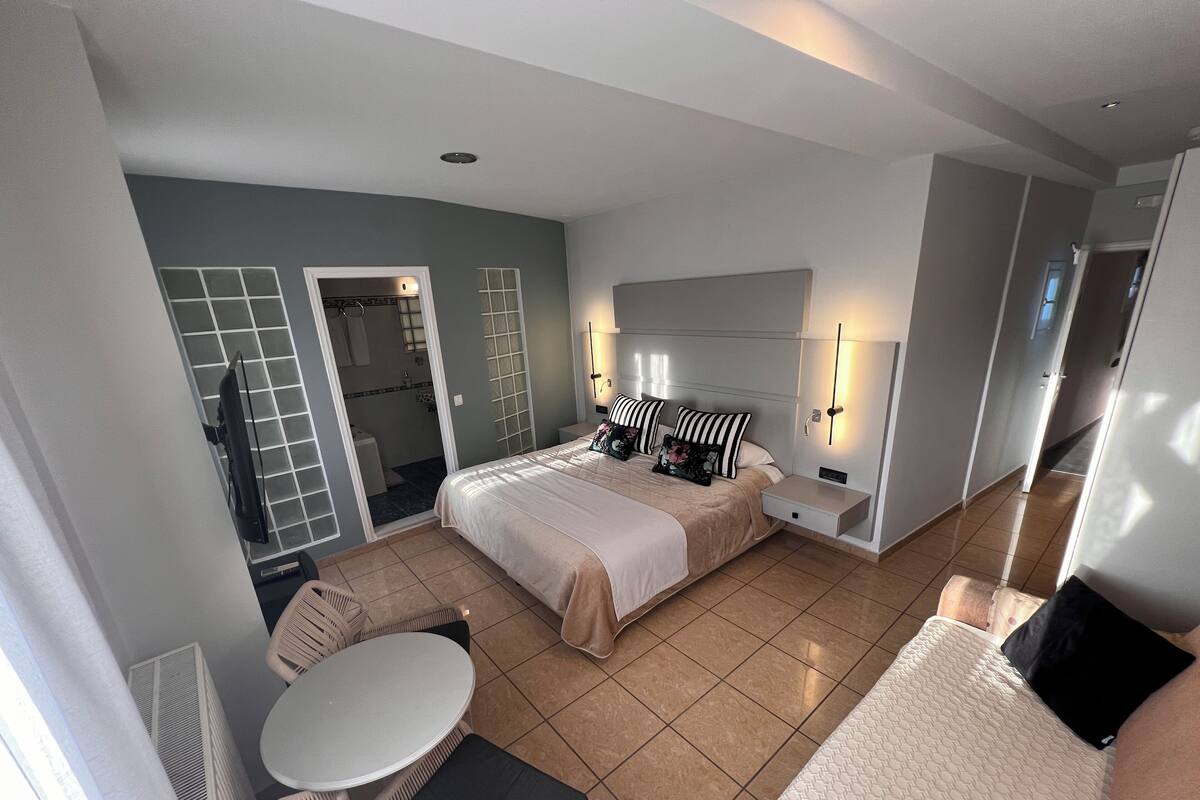 Een moderne hotelkamer met een groot bed, een kleine tafel en een bank. Een badkamer met douche en toilet is zichtbaar door een geopende deur.