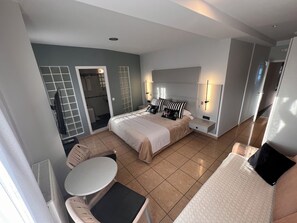 Ein modernes Hotelzimmer mit einem großen Bett, einem kleinen Tisch und einer Couch. Ein Badezimmer mit Dusche und Toilette ist durch eine offene Tür sichtbar.