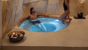 A man relaxing in a hot tub with a woman standing beside it, set against a rock wall. - Aressana Spa Hotel & Suites - Small Luxury Hotels of the World (Santorini)