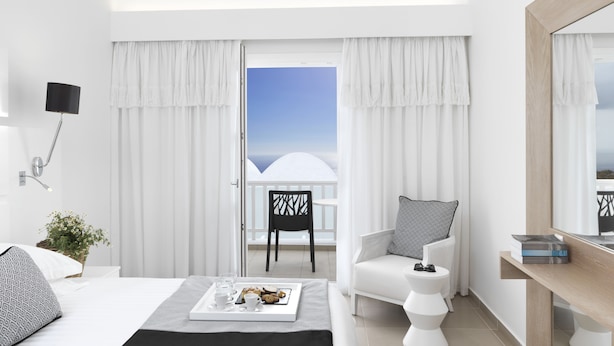 A modern hotel room with a bed, a sitting area with a chair and a small table, a balcony with a view, and a large mirror. - Aressana Spa Hotel & Suites - Small Luxury Hotels of the World (Santorini)