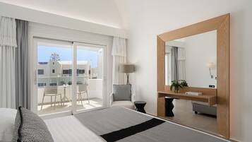 Habitación de hotel con cama, un espejo grande, escritorio con lámpara, una silla y balcón con vistas a edificios.
