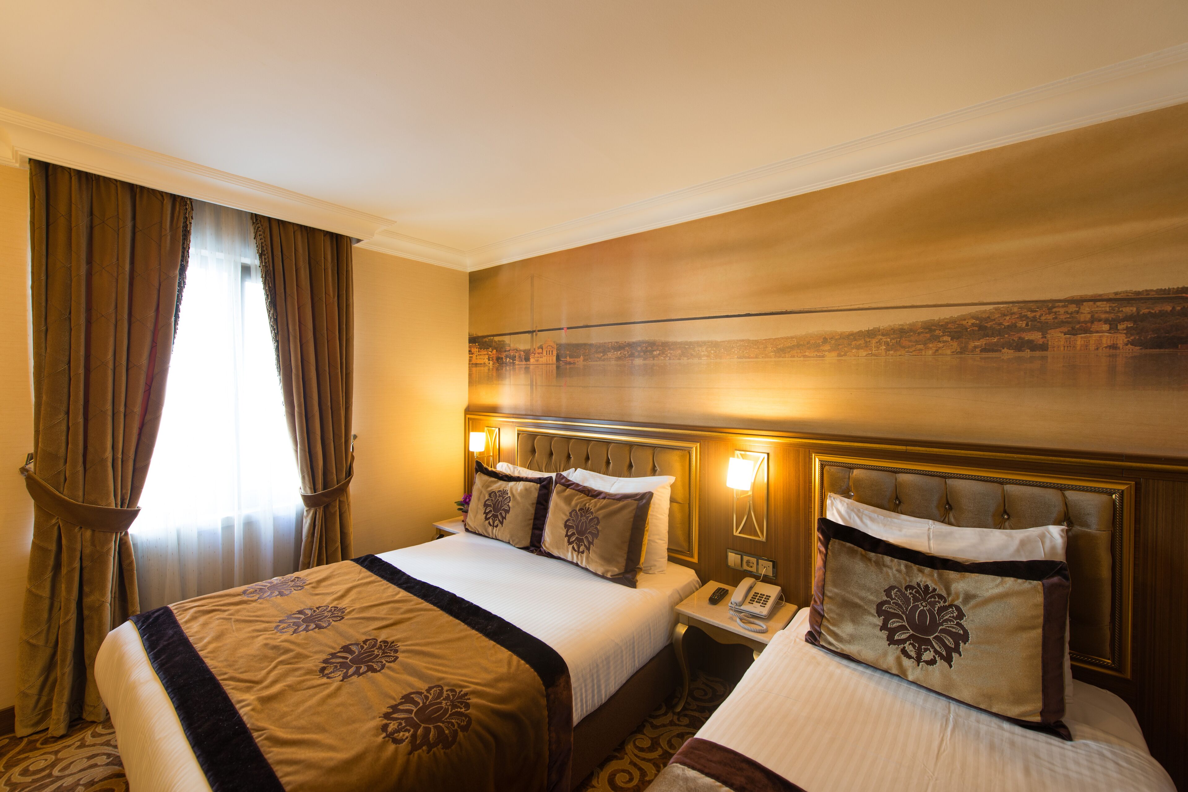 A hotel room with a large bed, a bedside table with a phone, and a view of a cityscape through the window.