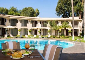 Un área de comedor junto a la piscina, con una mesa preparada para dos personas, rodeada de sillones y sombrillas, con un edificio al fondo que tiene balcones y una piscina.