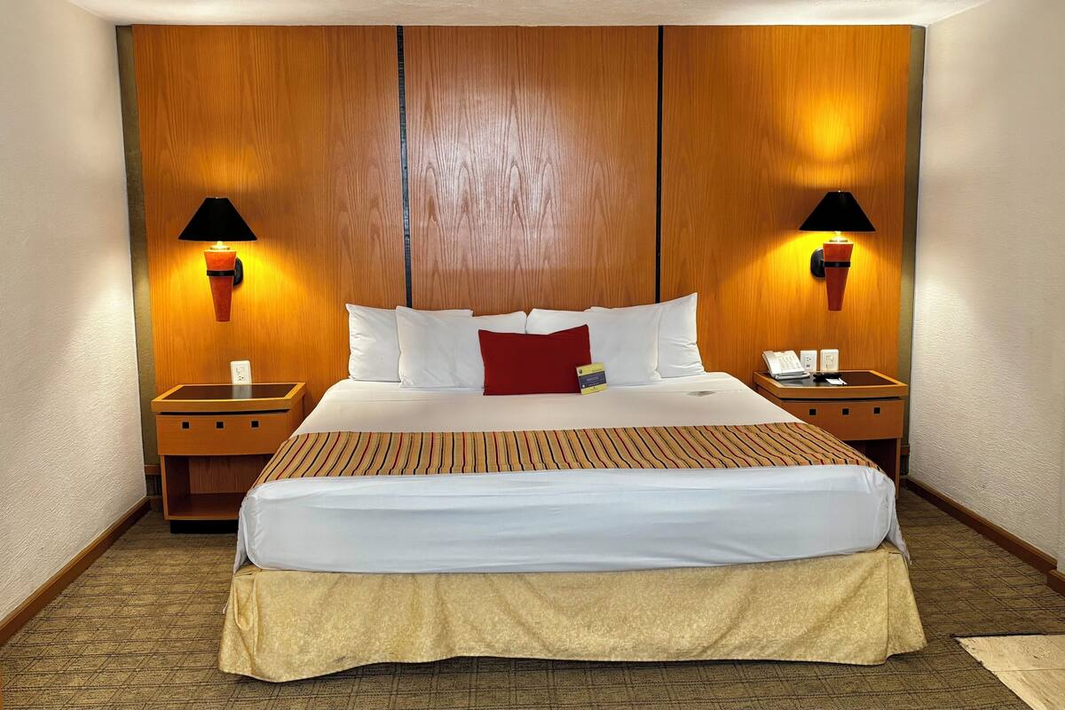 Une chambre d’hôtel avec un grand lit, une tête de lit en bois, deux lampes de chevet, une table de chevet avec un téléphone et une petite table de nuit.