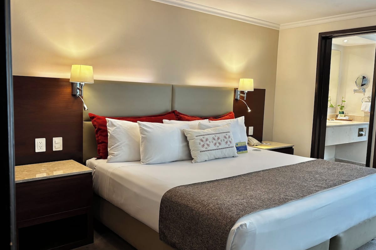Une chambre d’hôtel avec un lit, des lampes de chevet, une table de nuit, un miroir et une salle de bain visible à travers une porte ouverte.