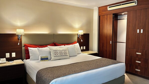 Una habitación de hotel con cama, mesitas de noche, escritorio, armario y unidad de aire acondicionado.