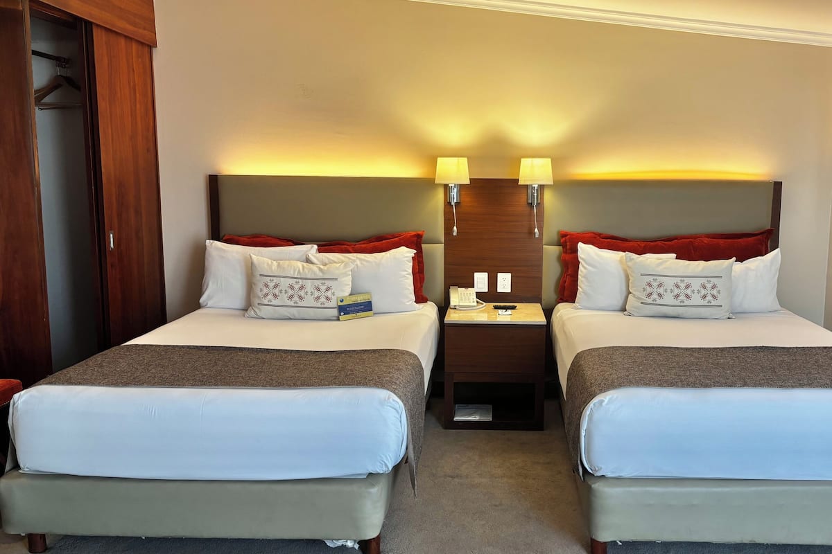 Une chambre d’hôtel avec deux lits, une table de chevet avec un téléphone et une télécommande, et une lampe fixée au mur.