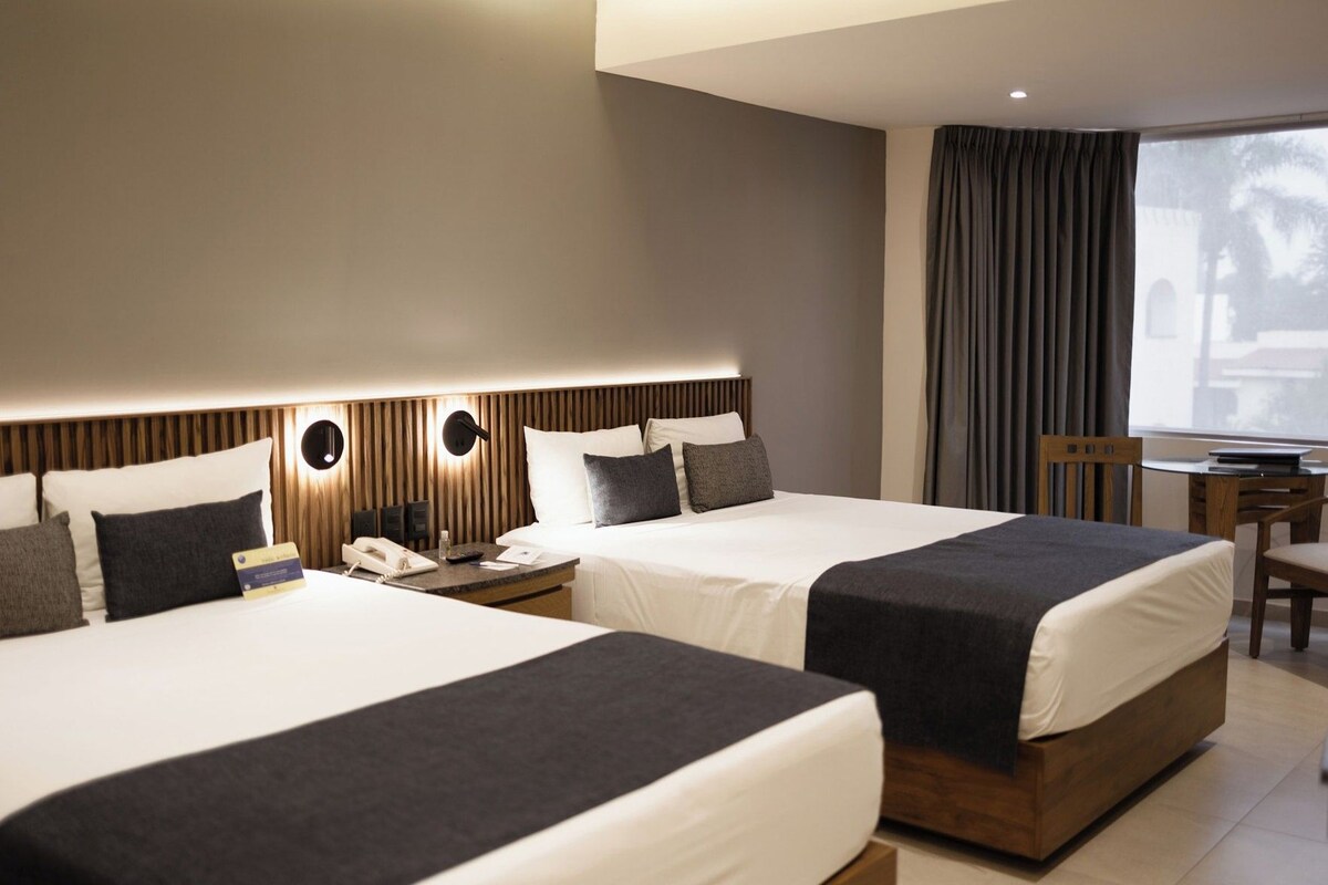 Habitación de hotel con dos camas, un escritorio y vistas al exterior.