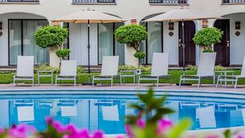 A poolside area with lounge chairs and umbrellas, a pool, and a building with balconies and windows.