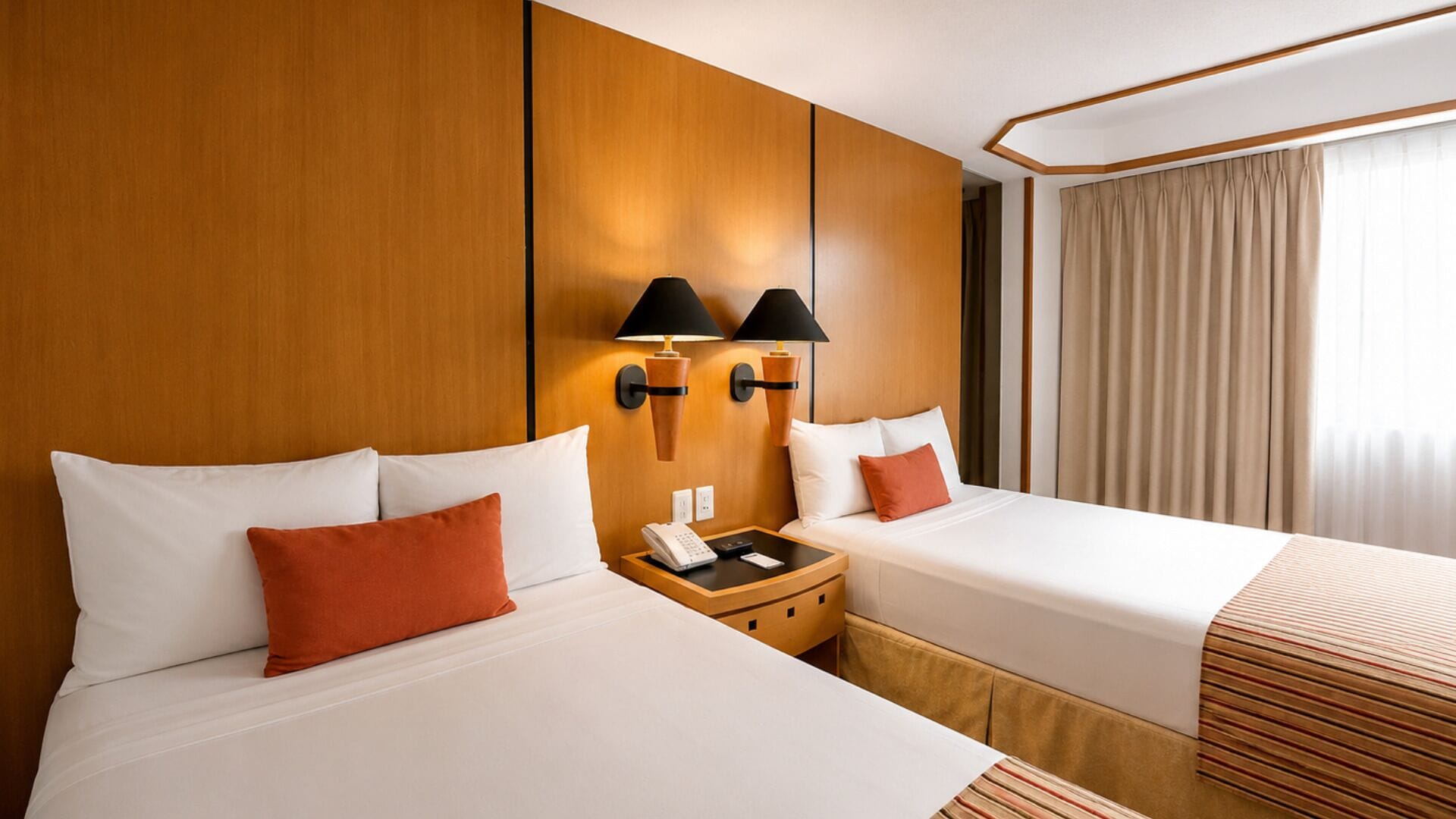 Une chambre d’hôtel avec deux lits, une tête de lit en bois, une table de chevet avec un téléphone et une lampe, et une fenêtre avec des rideaux.