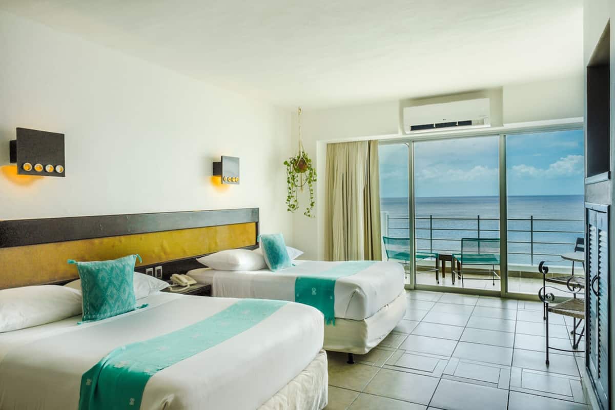 Une chambre d’hôtel avec deux lits, une grande fenêtre donnant sur la mer, un balcon avec une table et des chaises, et une plante suspendue.