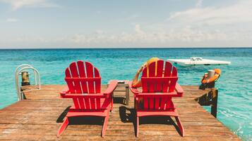 Two red Adirondack chairs on a wooden pier with a view of the ocean and a boat in the distance.