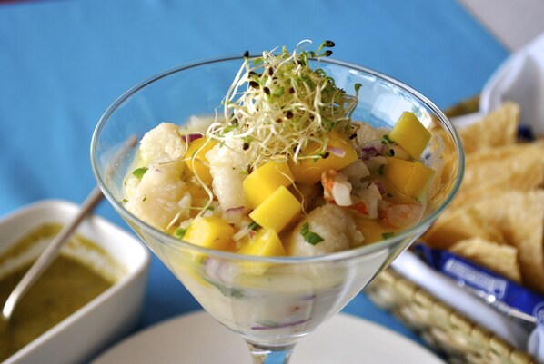A clear glass bowl filled with a seafood salad, garnished with sprouts, accompanied by a side of dipping sauce and crackers. - Hotel B Cozumel (Cozumel)