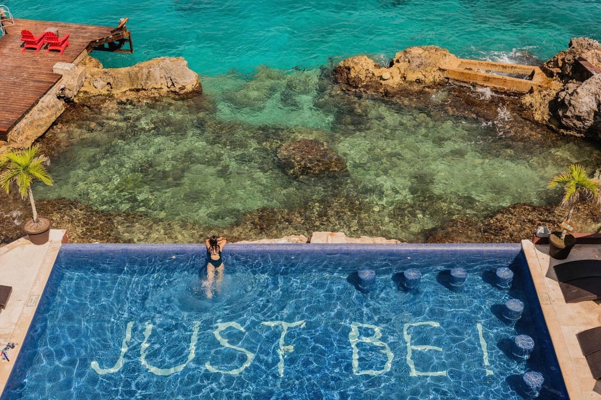 A pool with the words 'JUST BE!' written on the edge, surrounded by clear water and rocks.