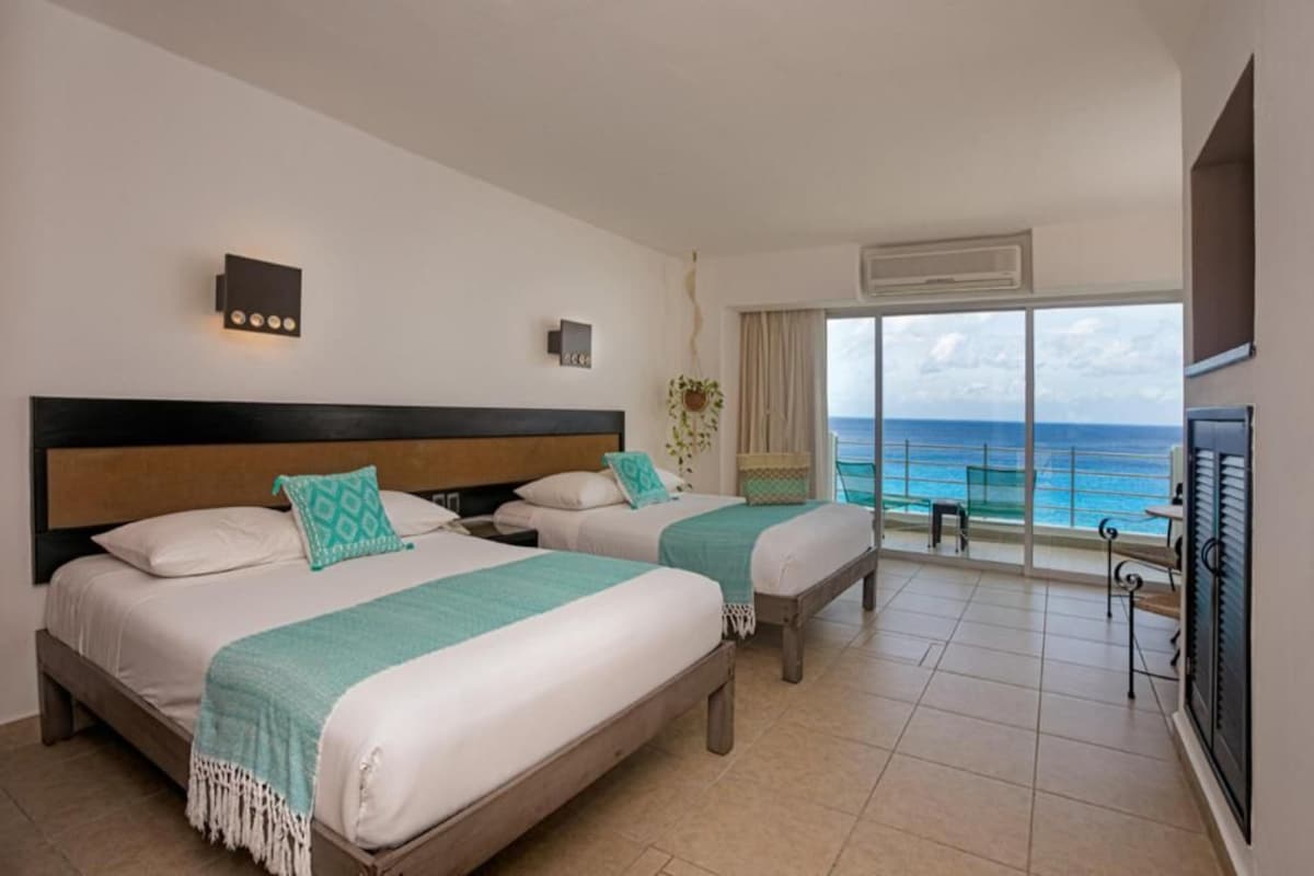 Habitación de hotel con dos camas, un ventanal con vistas al océano, un televisor y una planta.