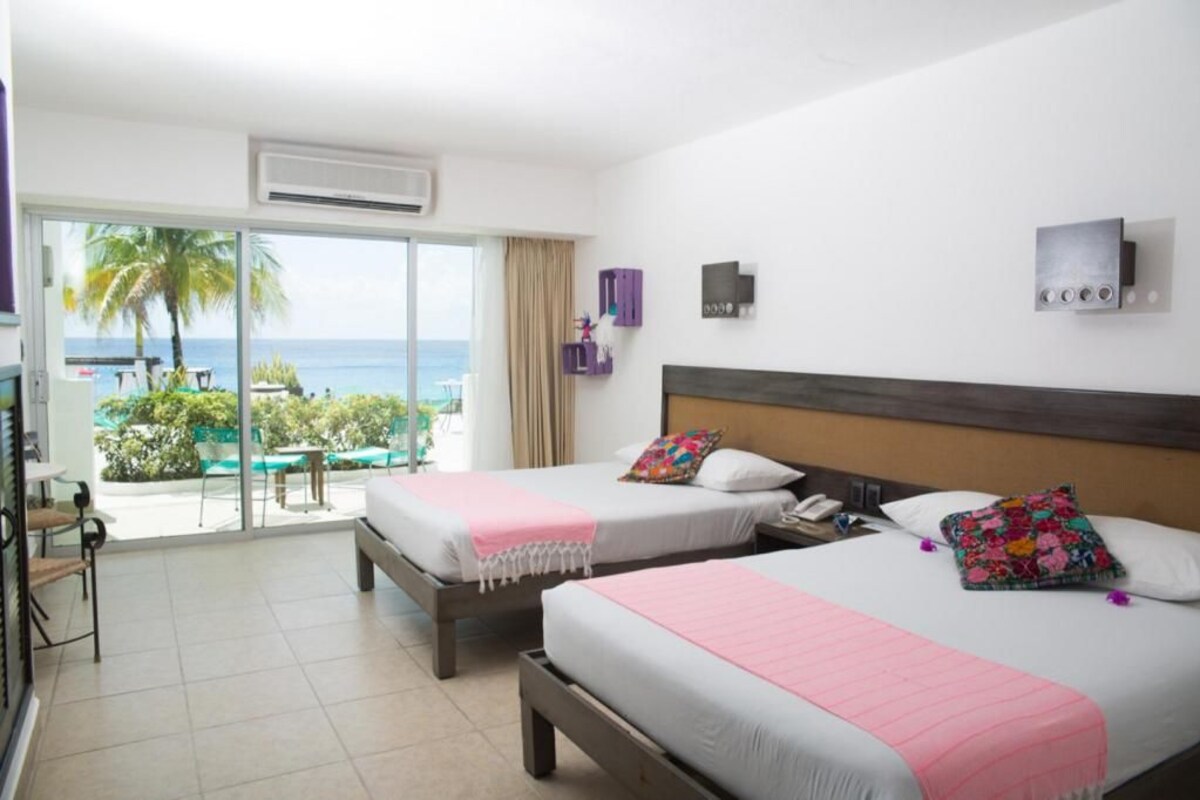 Habitación de hotel con dos camas, un balcón con vista al mar y una pequeña mesa con sillas.