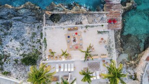 Vista aérea de un complejo turístico frente a la playa, con un patio, sillas de descanso y palmeras.