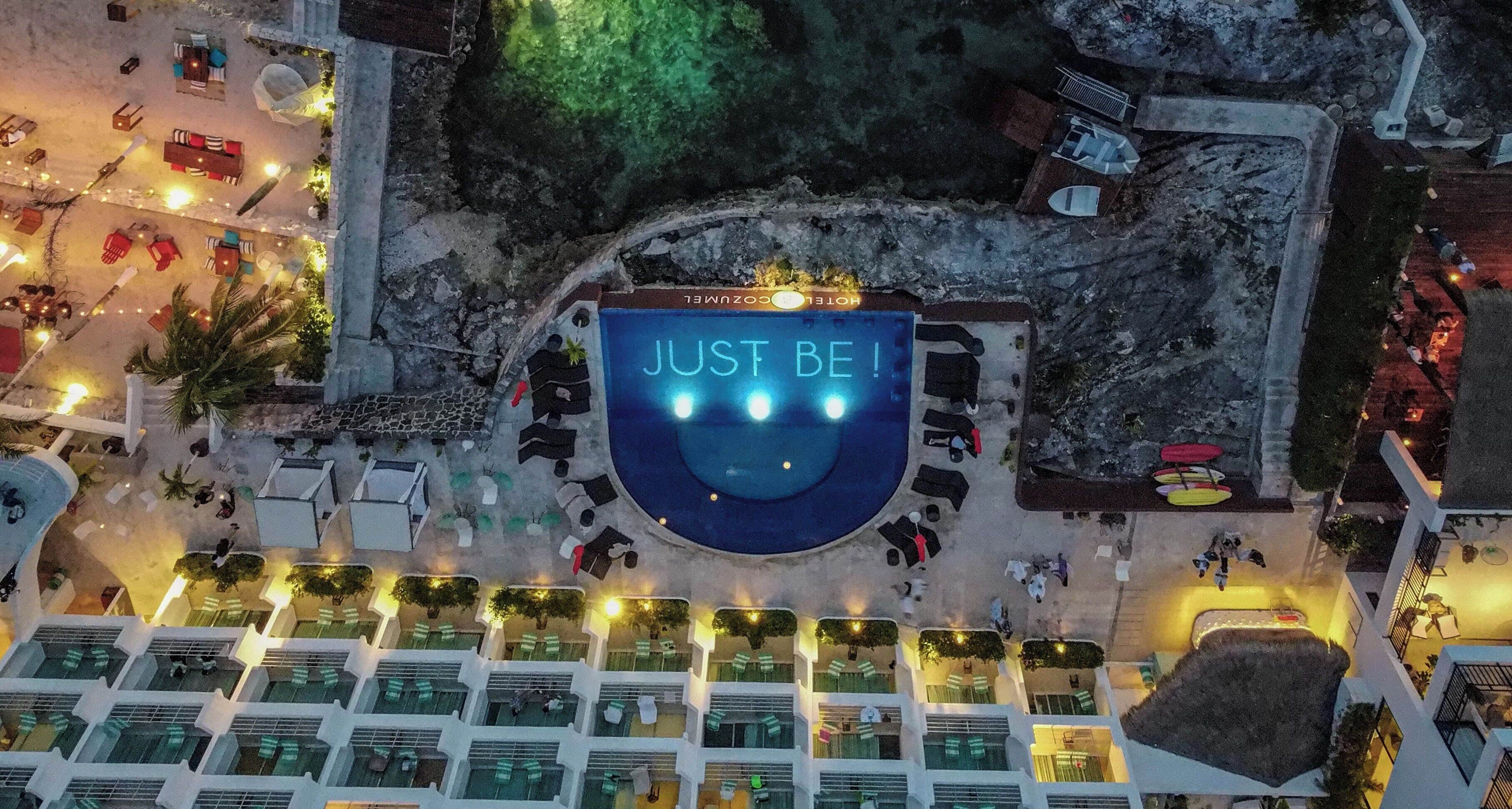 Vue aérienne d’un hôtel avec une piscine, un espace de détente en plein air et un panneau sur lequel on peut lire : « SOYEZ VOUS-MÊME ! ».