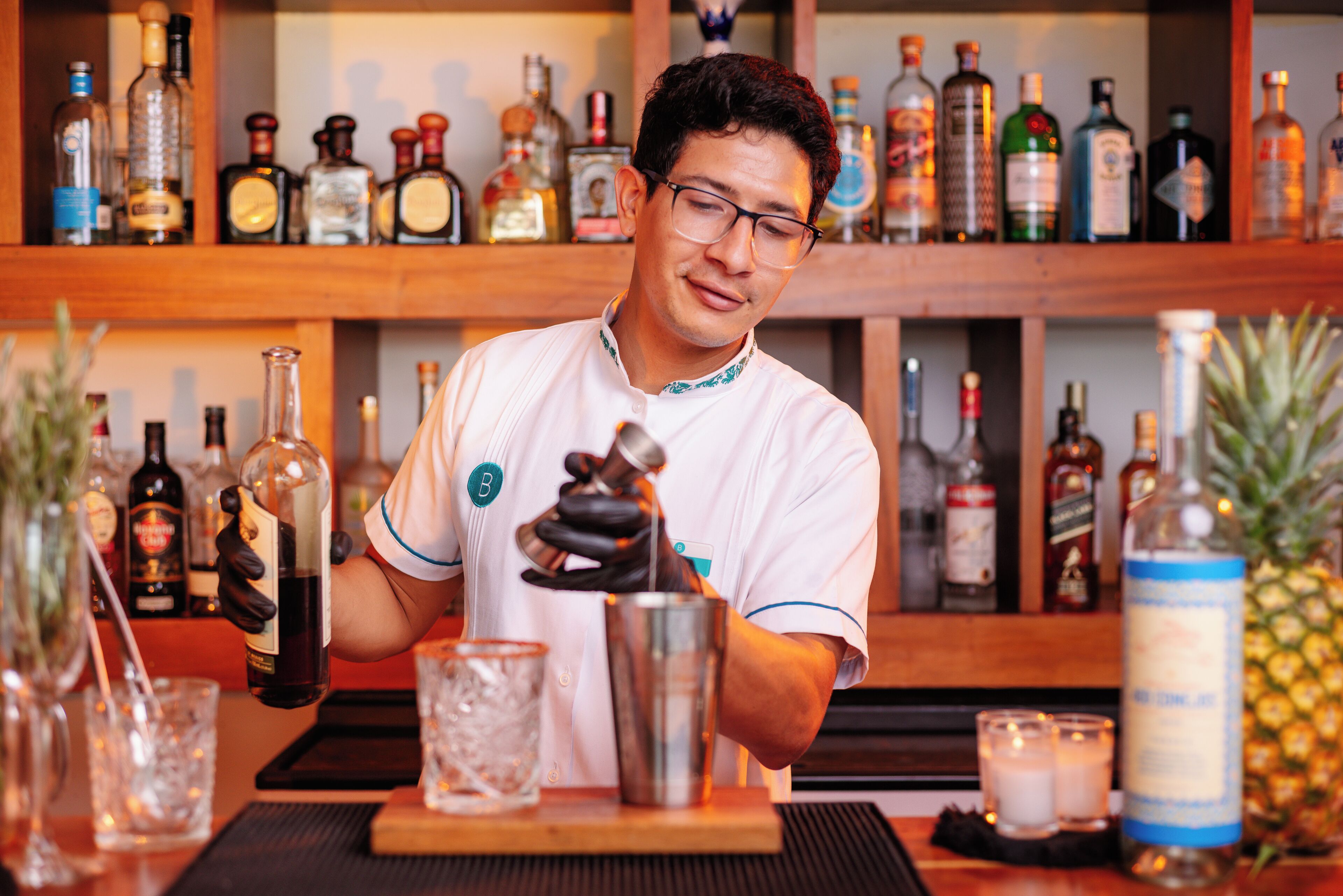 Ein Barkeeper bereitet einen Drink an der Bar zu, im Hintergrund sind verschiedene Flaschen und Gläser zu sehen.