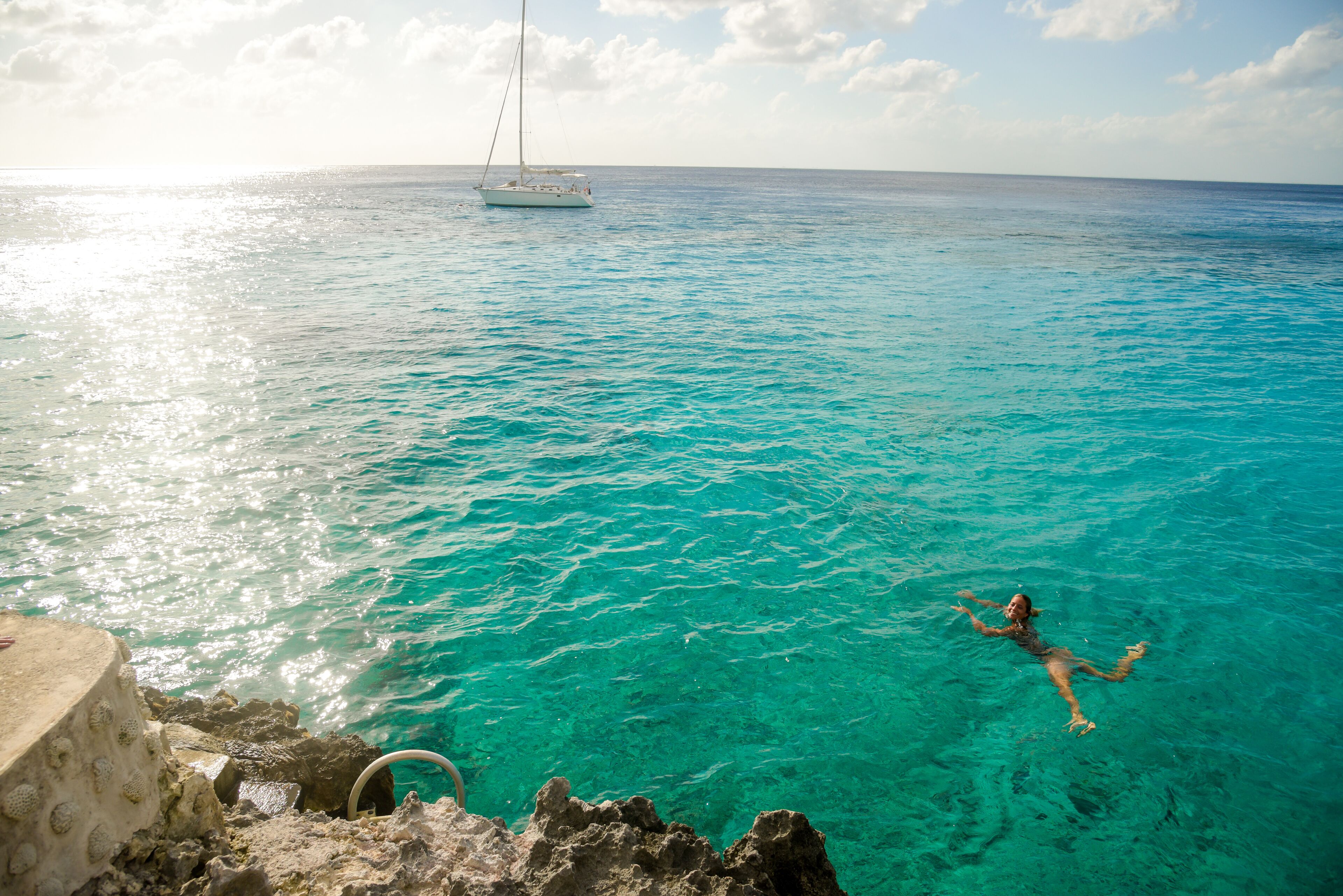 A person swimming in clear turquoise water with a sailboat in the distance.