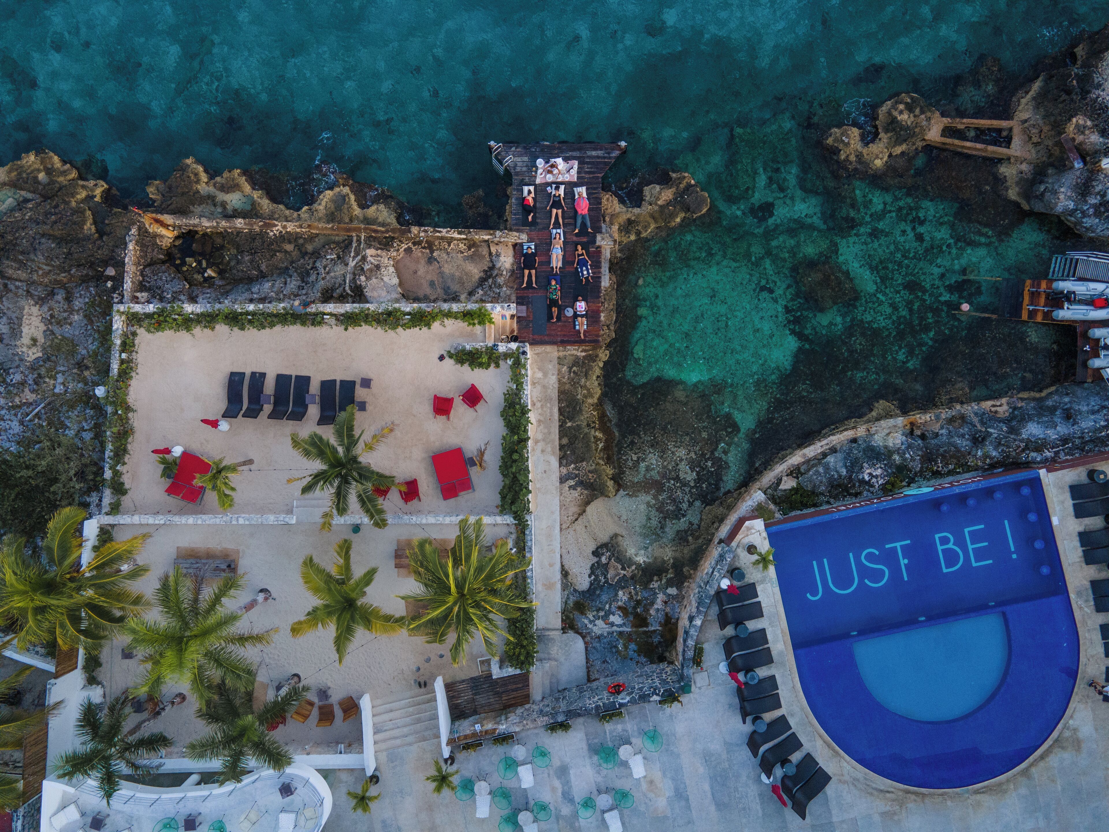 Vista aerea di un resort sulla spiaggia, con area piscina, lettini e palme.