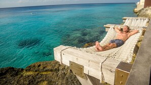 Una persona descansando en una hamaca al borde de un acantilado con vistas a aguas turquesas.