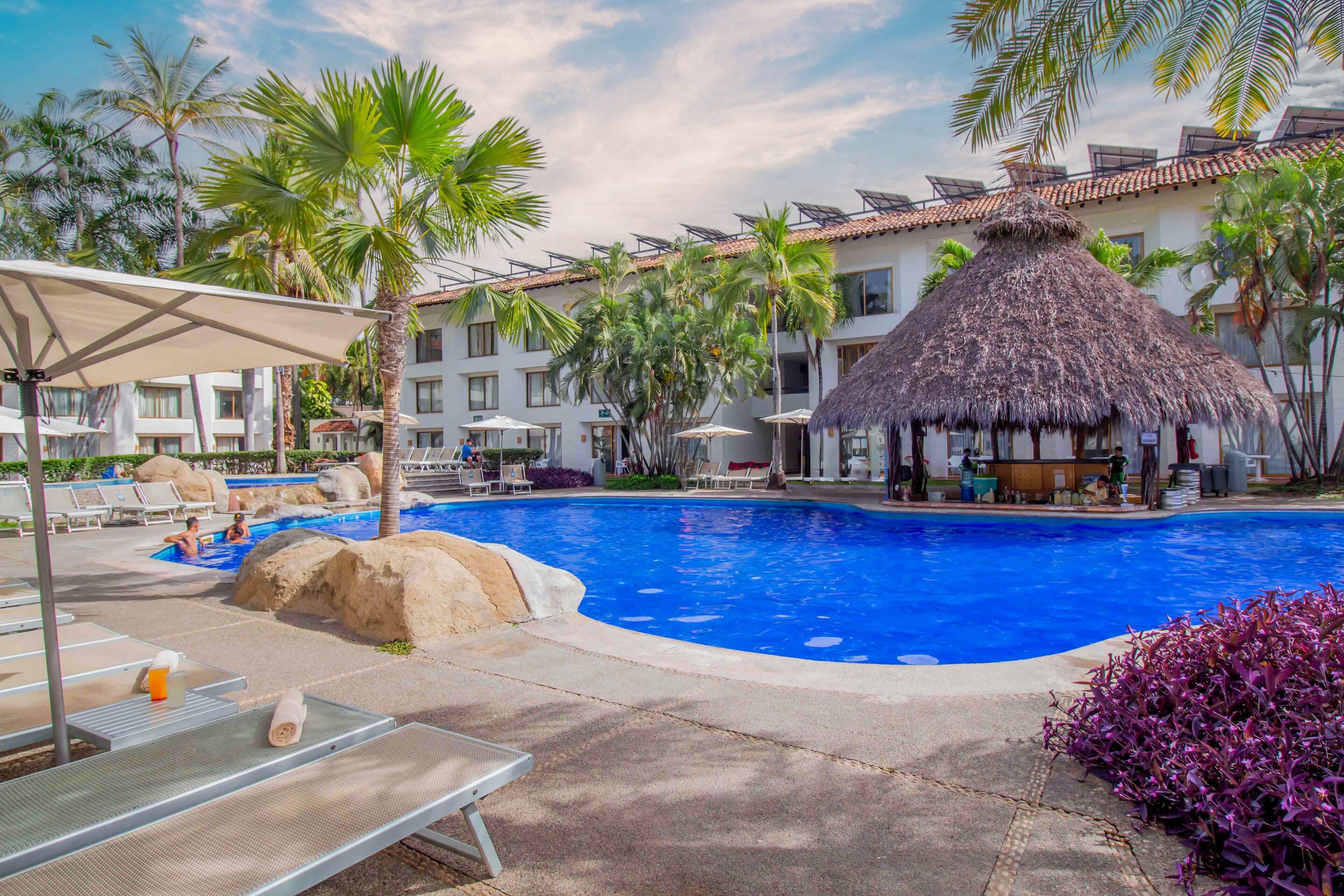 A swimming pool with lounge chairs and umbrellas, surrounded by palm trees and a building with a thatched roof.