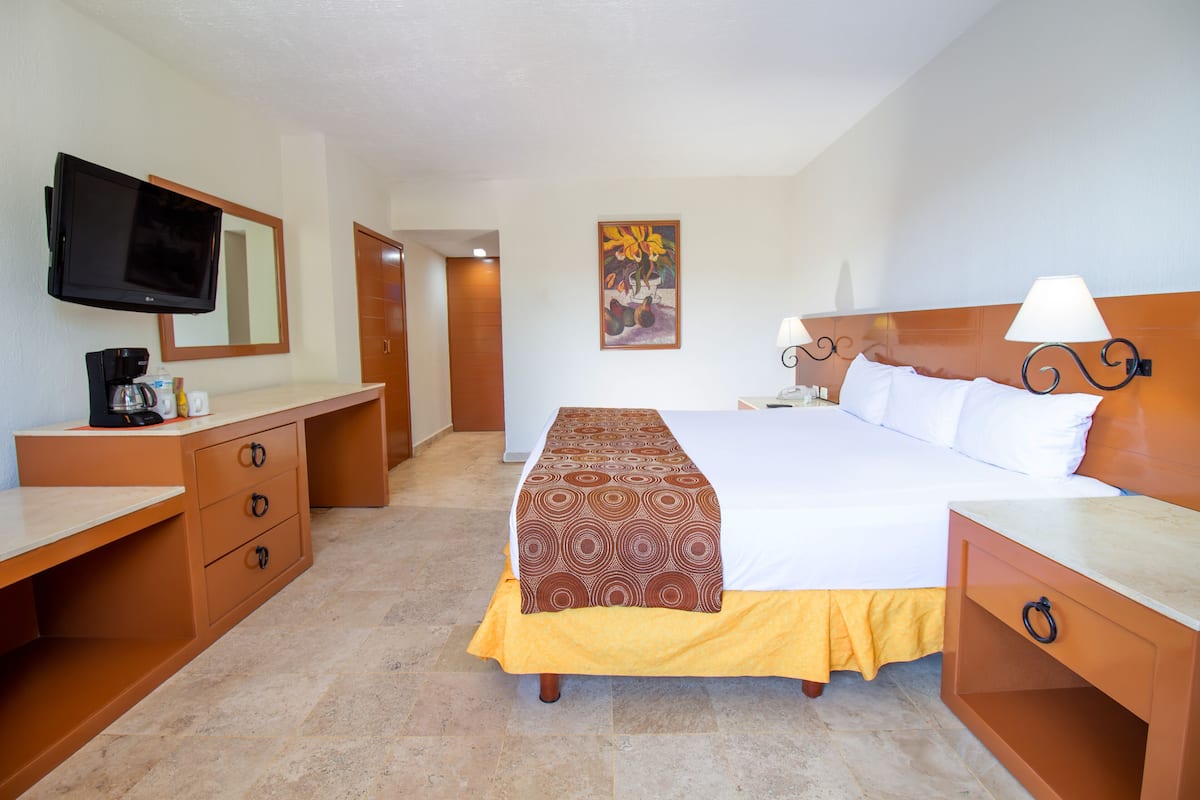 Une chambre d’hôtel avec un lit, des tables de chevet, un bureau avec une chaise, une télévision et un tableau au mur.