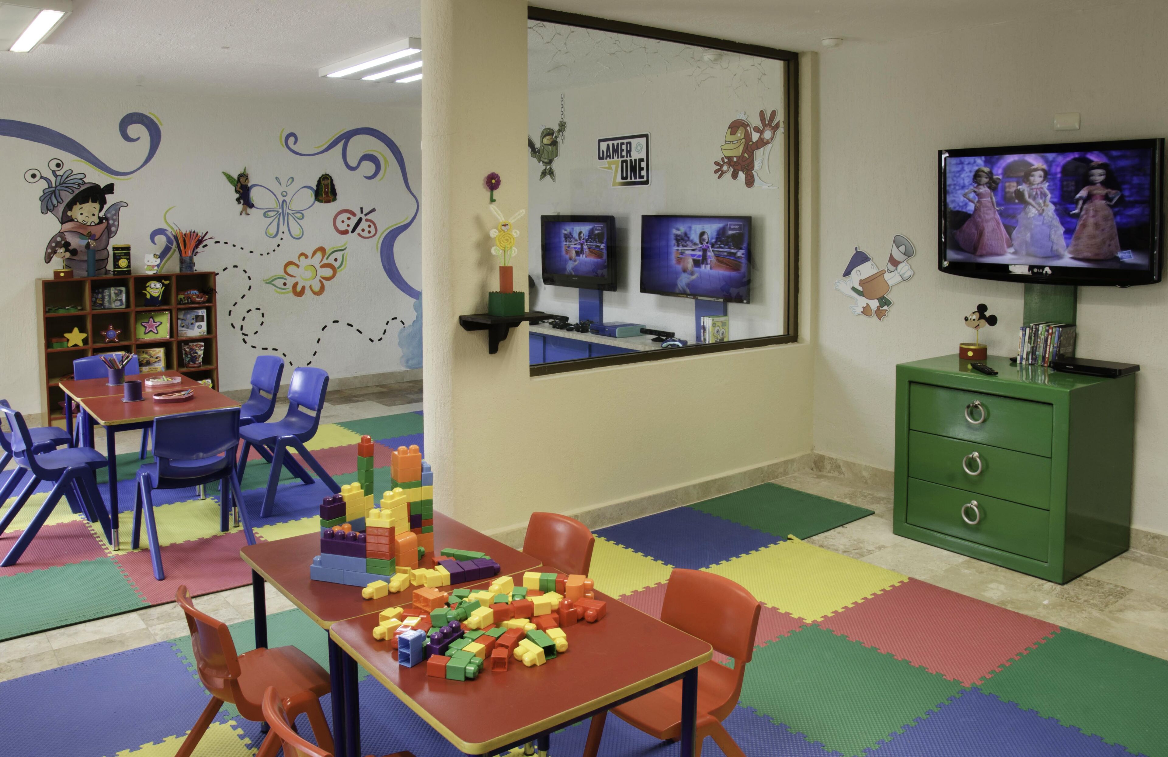 Un área de juegos infantiles con tapetes de colores, una mesa con bloques de construcción y dos televisores en la pared.