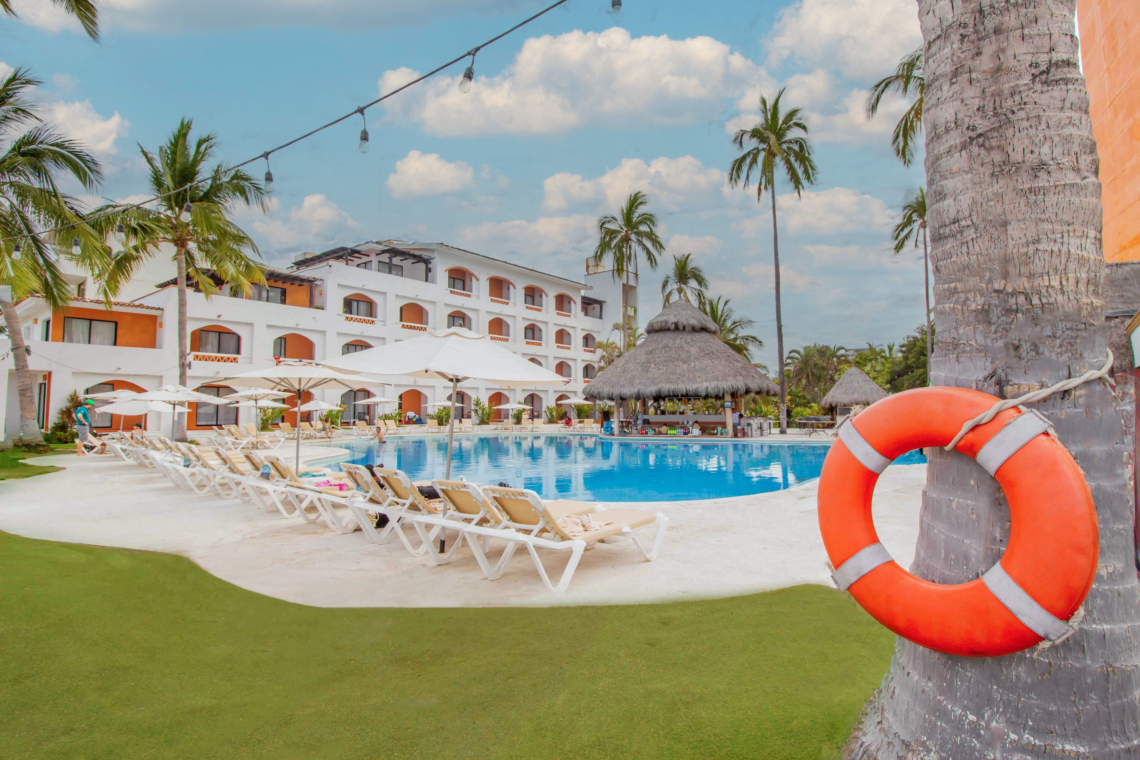 A pool area with lounge chairs, a lifebuoy, palm trees, and a building with multiple balconies.