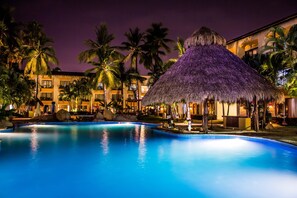 A swimming pool with a thatched-roof structure, palm trees, and a building with balconies. - Plaza Pelicanos Club Beach Resort All Inclusive (Puerto Vallarta)