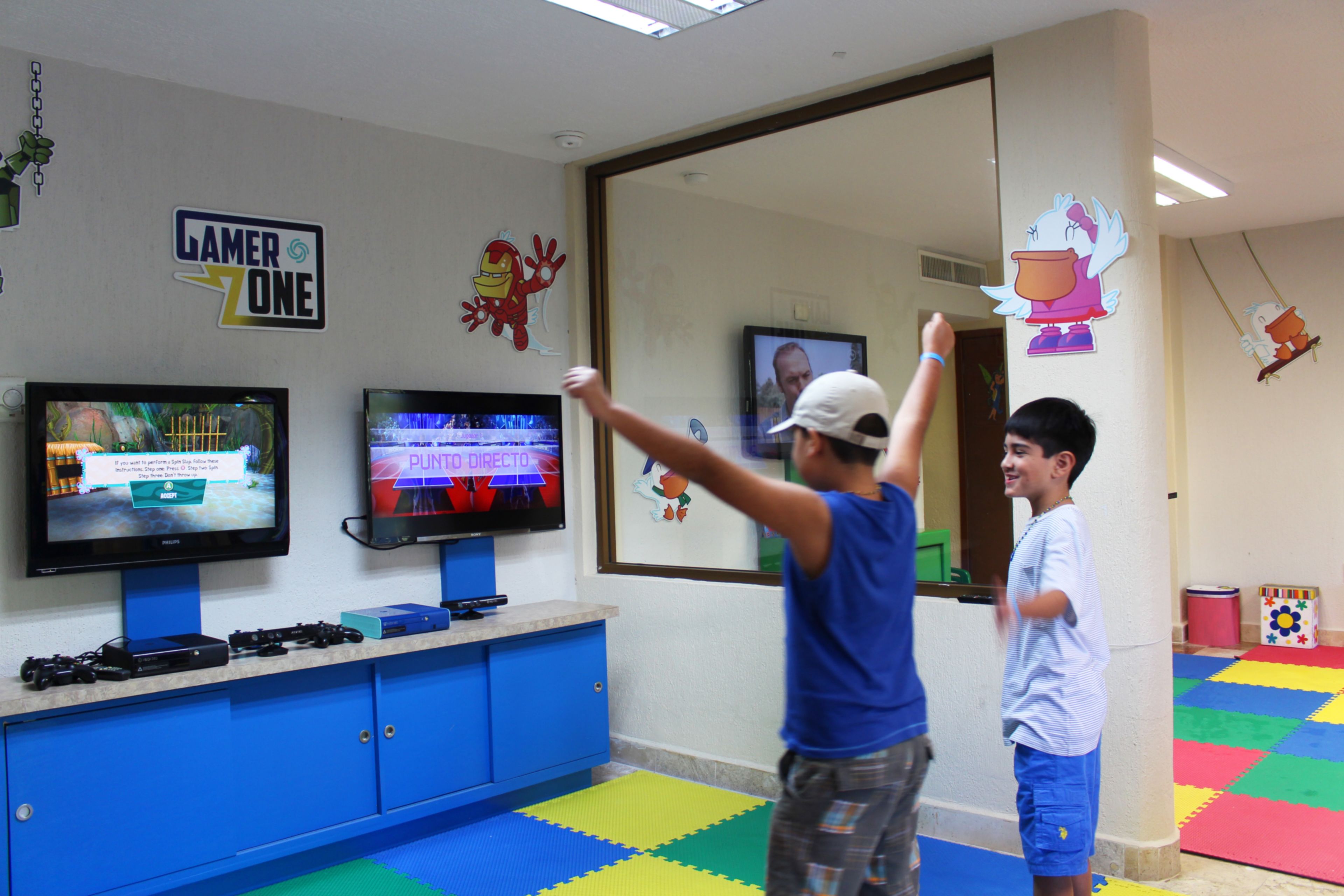 Habitación con consolas de videojuegos, dos televisores mostrando juegos y alfombras de juego coloridas en el suelo.