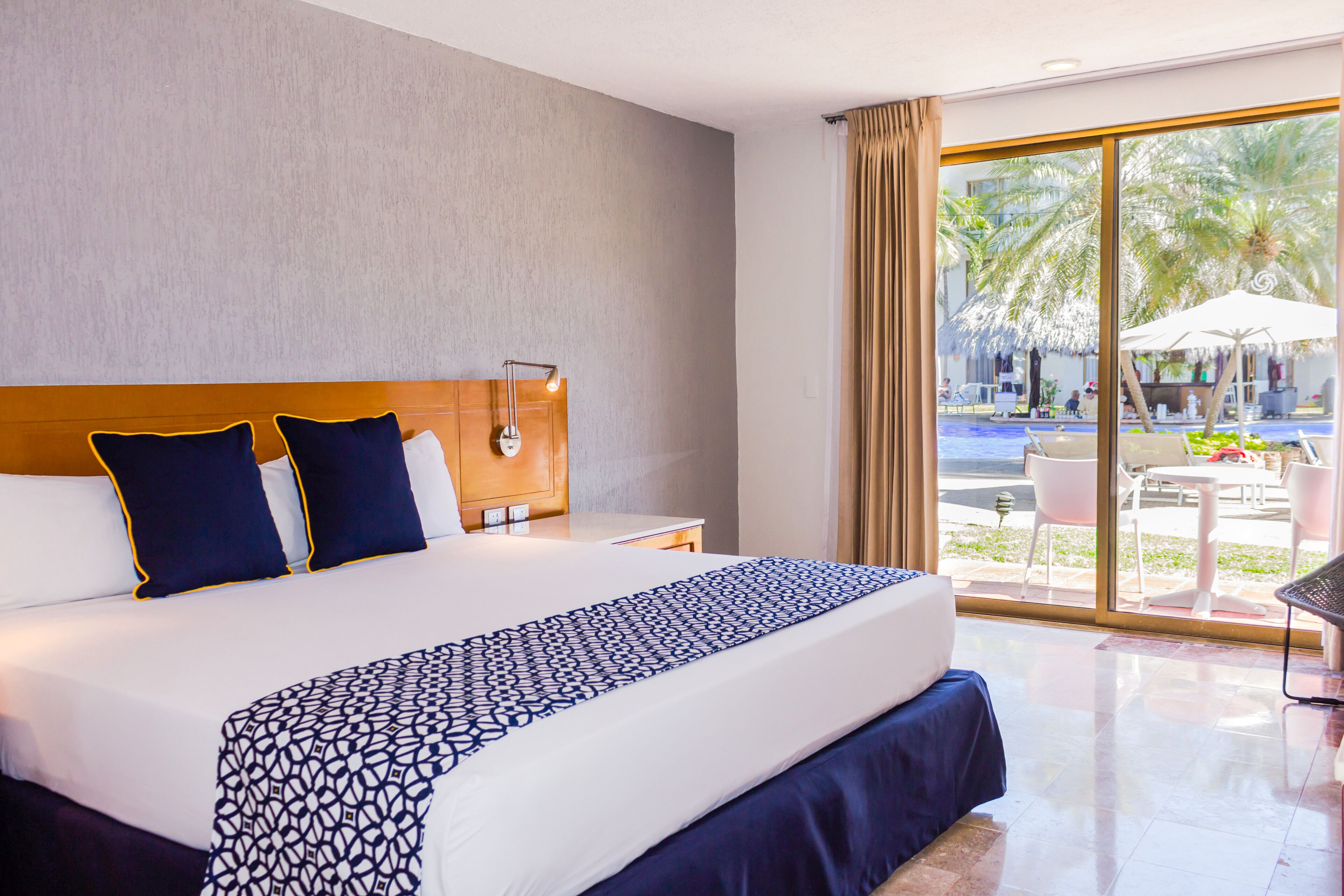 A hotel room with a large bed, a nightstand, a desk, and a view of an outdoor pool area.