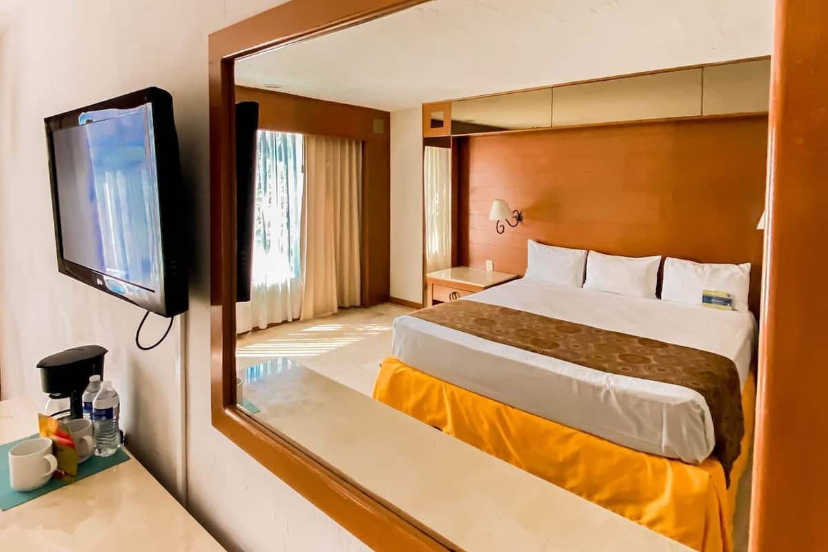 Une chambre d’hôtel avec un lit, une télévision, un miroir et une table sur laquelle se trouvent une tasse et une bouteille d’eau.