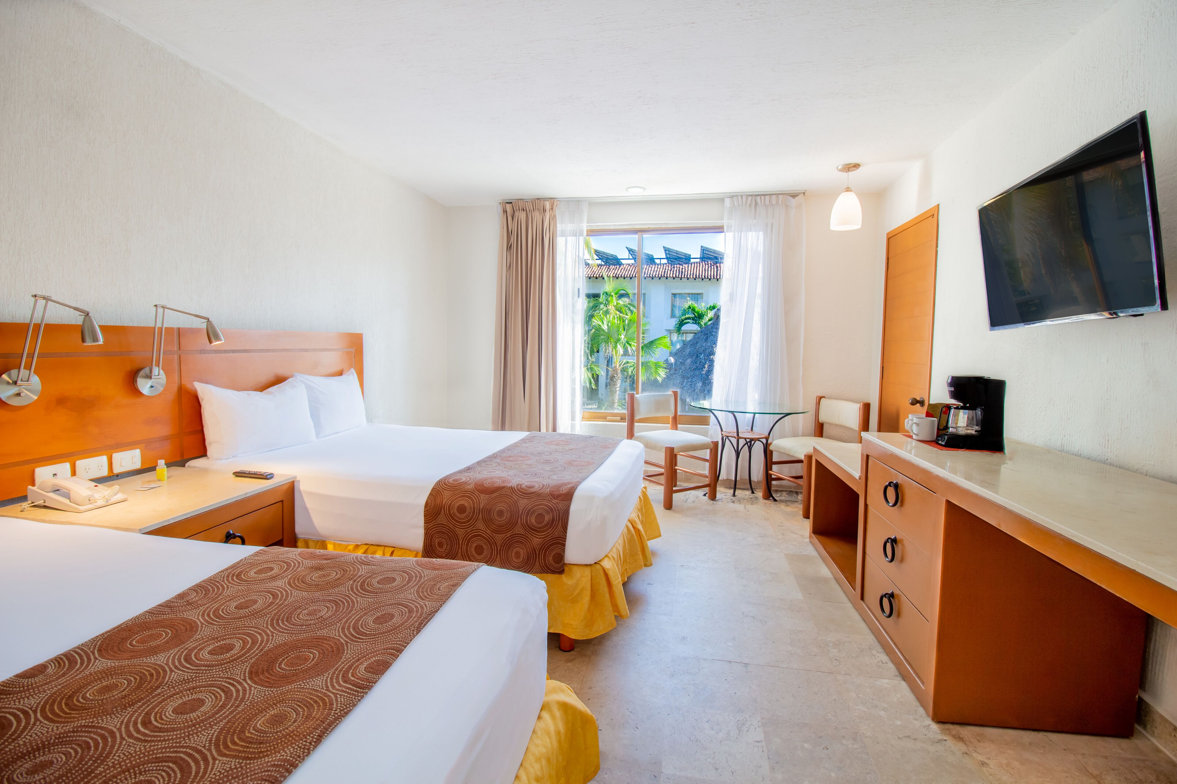 Une chambre d’hôtel avec deux lits, un bureau, une chaise, une télévision et une fenêtre donnant sur des palmiers.