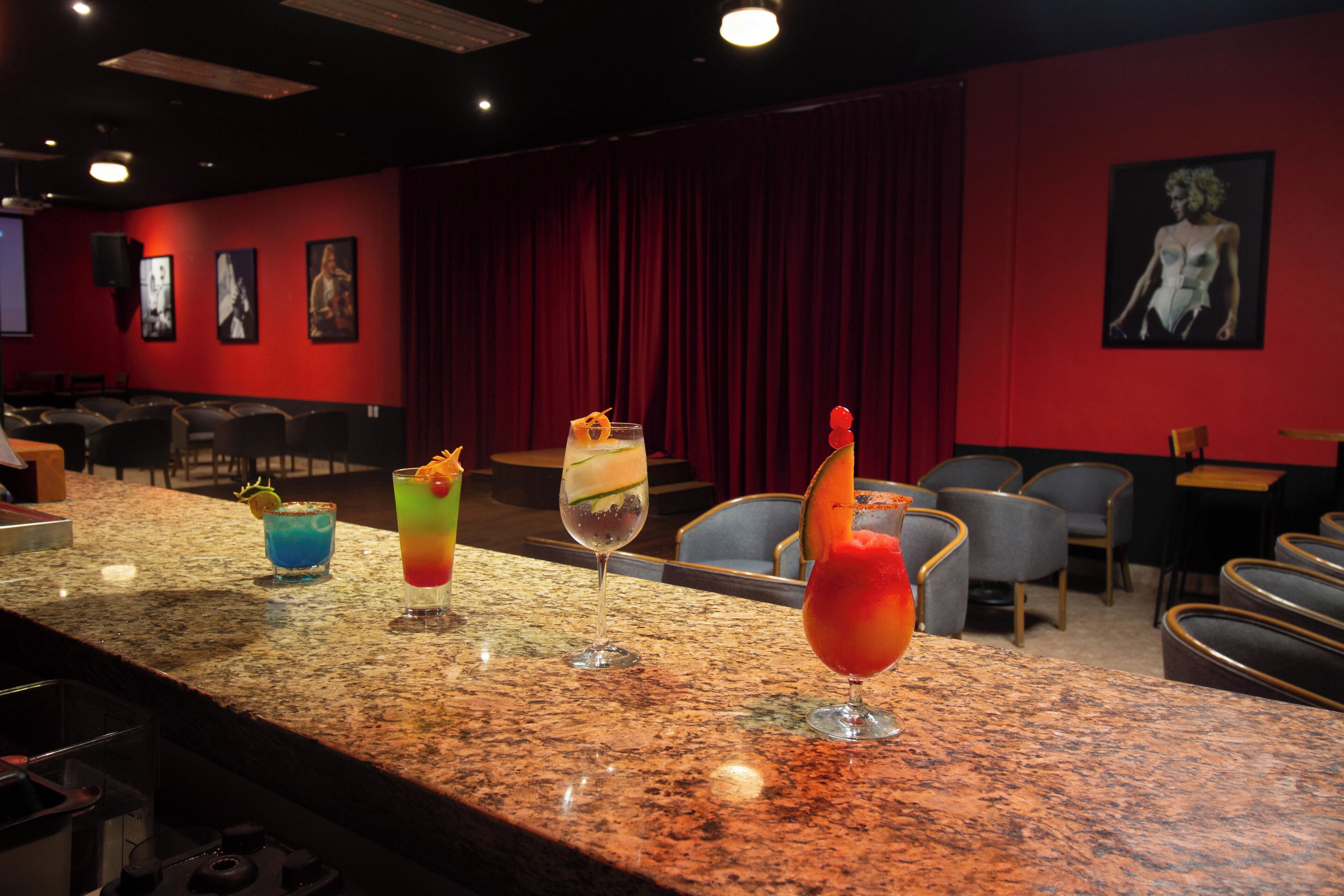 Zona de bar amb un taulell de granat que mostra diferents còctels de colors, un fons de cortina vermella i una zona de seients amb cadires.