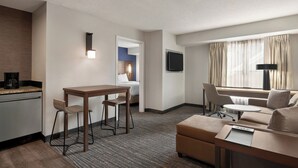 Ein Hotelzimmer mit Küchenzeile, einem Essbereich inklusive Tisch und Stühlen, einer Couch, einem an der Wand befestigten Fernseher und einem Fenster mit Vorhängen.