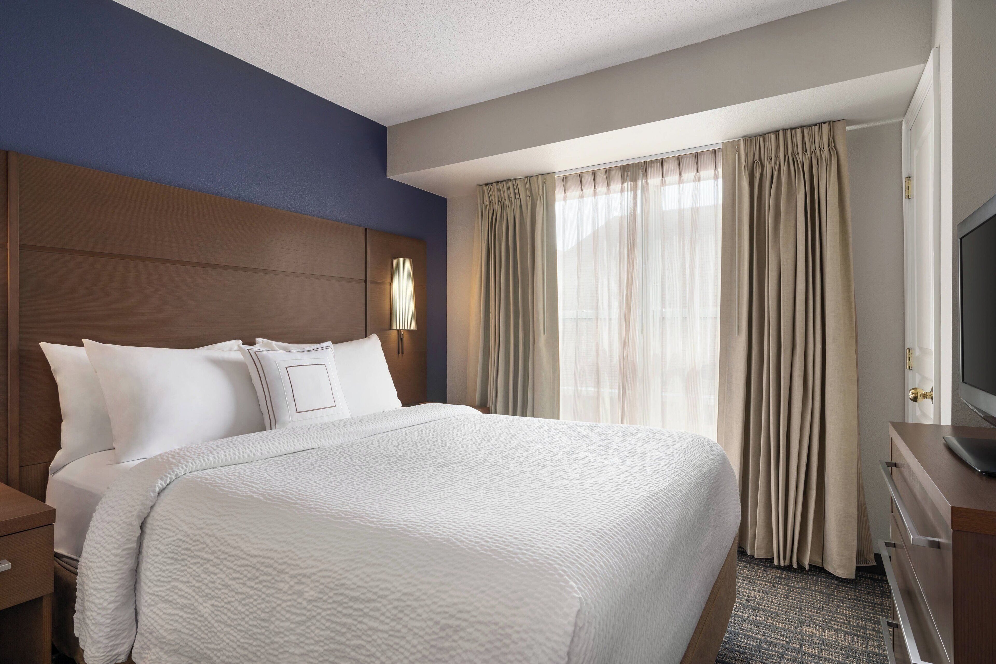 A hotel room with a large bed, a wooden headboard, a nightstand, a TV, and a window with curtains.