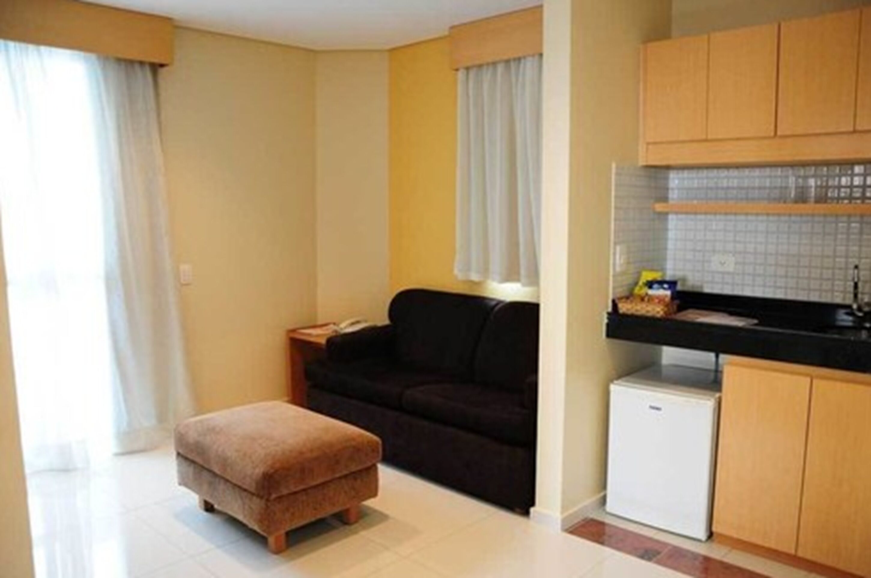 A compact living space with a kitchenette, a sofa, a small ottoman, and a refrigerator.