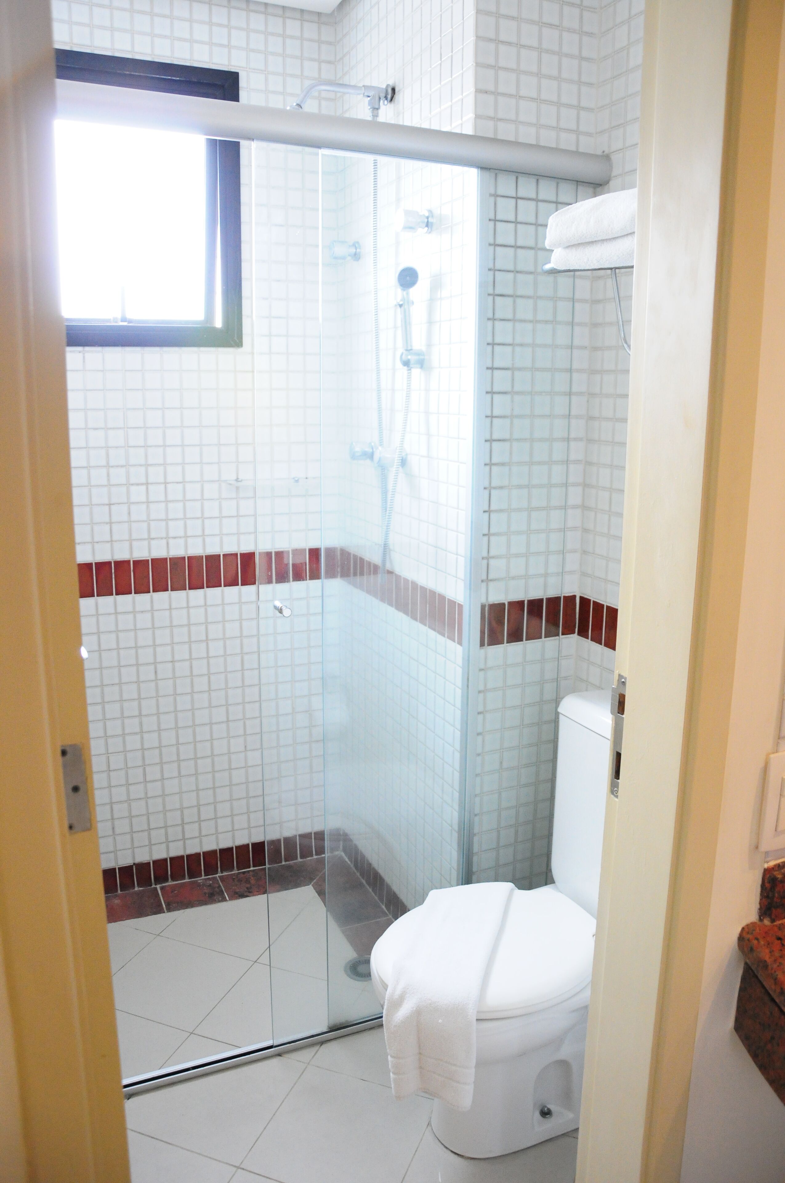 A bathroom with a glass-enclosed shower, white tiles, and a red-tiled accent strip.